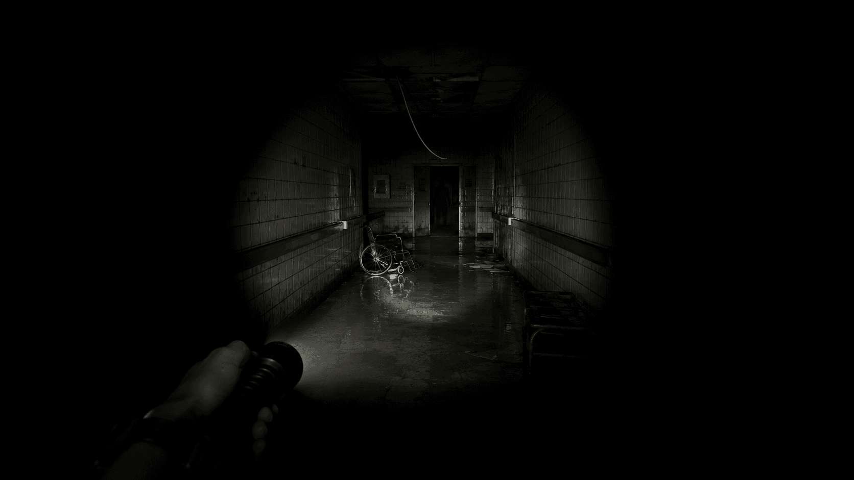 A dark hospital corridor seen from a first-person view, lit only by a narrow torch beam picking out a wheelchair and a slumped shape further down.